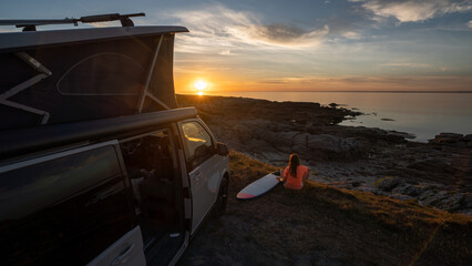 Surfer girl sitting near her mini van and looking on the ocean at summer sunset with a surfboard on her side © AA+W