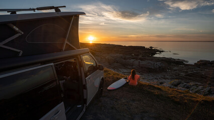 Surfer girl sitting near her mini van and looking on the ocean at summer sunset with a surfboard on...