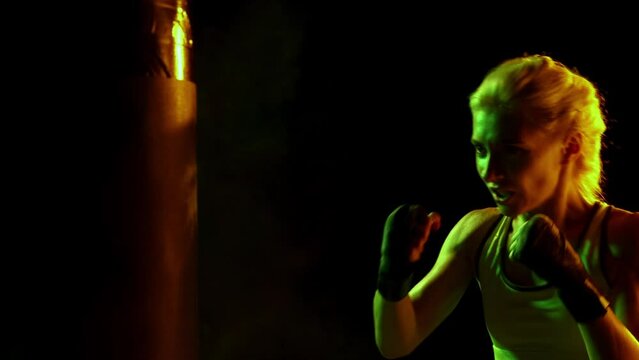 Sportive woman, female professional boxer practices punch on punching bag in dark smoky gym in neon light. Sport, action, power, energy concept.