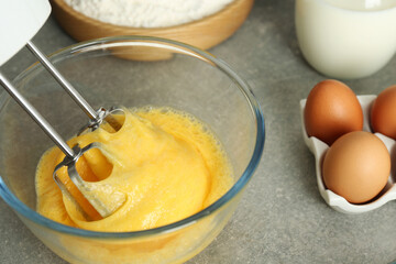 Beating eggs in glass bowl with mixer on light grey table, closeup