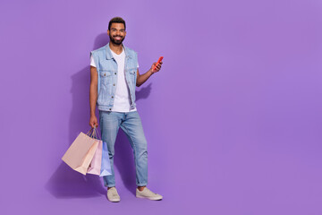 Full length photo of pretty funny man wear jeans waistcoat holding shoppers modern device empty space isolated violet color background