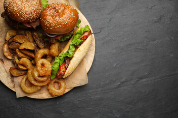 Tasty burgers, hot dog, potato wedges and fried onion rings on black table, top view with space for...