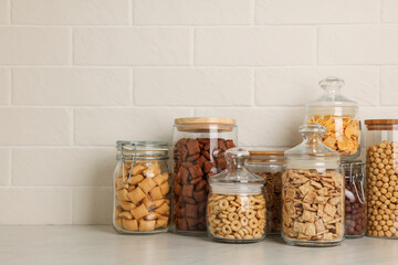 Glass containers with different breakfast cereals on white countertop near brick wall. Space for text