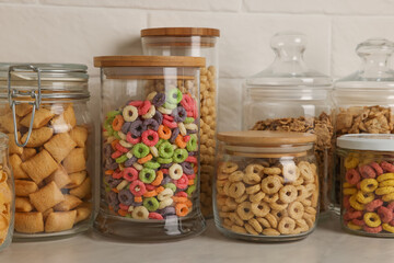 Glass containers with different breakfast cereals on white countertop near brick wall