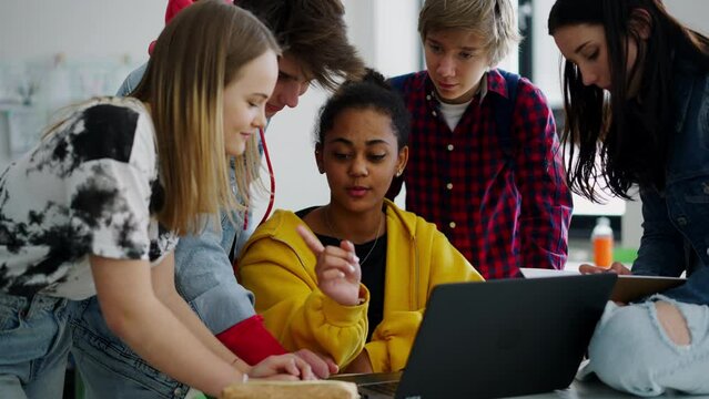 High school students sitting together at desk and using laptop and talking during break in classroom.