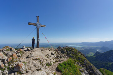 On top, young woman standing on the summit cross of Aggenstein in Austria and looking into the distance.