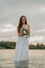 Fototapeta na wymiar Caucasian girl with black hair and holding a grass wreath with wildflowers and standing against the background of the sunset in the river. They practiced magic on the day of the Midsummer. 