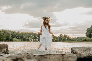 Cheerful girl at sunset is standing in shallow river in long white dress and with wreath on head and having fun. Woman splashes water with foot and laughs. On holiday of Ivan Kupala girls are guessing