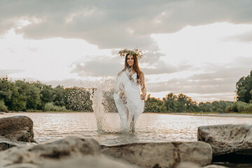 Cheerful girl at sunset is standing in shallow river in long white dress and with wreath on head...