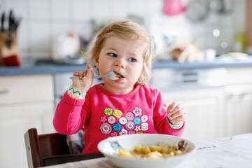 Adorable baby girl eating from fork vegetables and pasta. food, child, feeding and development concept. Cute toddler, daughter with spoon sitting in highchair and learning to eat by itself.