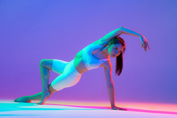 Studio shot of young flexible girl in fitness sport uniform practicing isolated on gradient pink-purple background in neon light. Sport, beauty, ad