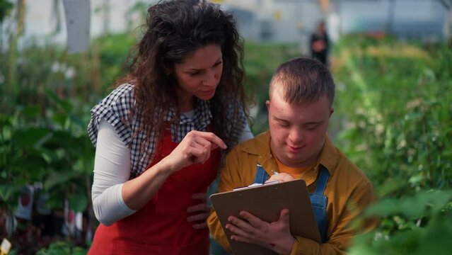 Experienced woman florist helping young employee with Down syndrome to check flowers on tablet in garden centre.