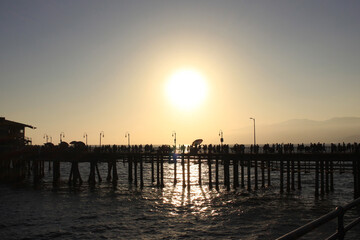 Backlit silhouettes of many unrecognizable people walking across the Santa Monica Pier at a slightly misty sunset with the sparkling sea