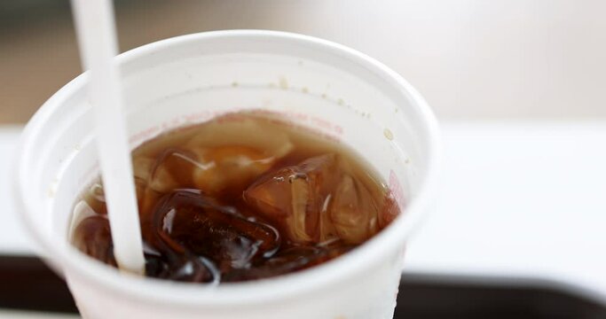 Hand holding straw and siring ice cola carbonate soft drink in white plastic glass.
