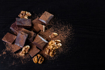 A pile of dark broken chocolate sprinkled with cocoa powder and walnuts on a dark background. Copy space