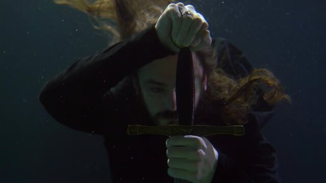 a long-haired man runs hand over the hilt of a sword in the dark water. close-up