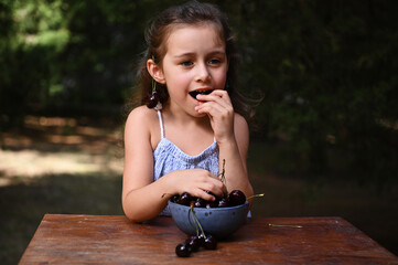 Beautiful baby girl, wearing a pair of ripe cherries earrings on her ears, eats ripe cherry berries in the summer garden