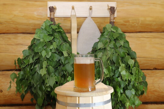 A mug of light beer stands on a wooden barrel on the background of fresh birch brooms hanging on a log wall.	