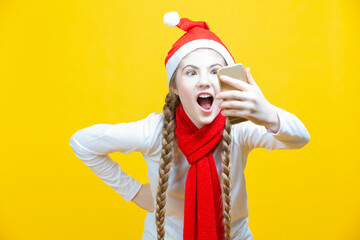 Funny Winsome Caucasian Teenager Holding Cellphone Taking Selfie While Smiling and Having Good Time And Making Faces in Christmas Santa Hat Posing Against Yellow Background.