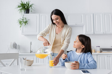 Obraz na płótnie Canvas cheerful babysitter pouring orange juice near child having breakfast in kitchen.
