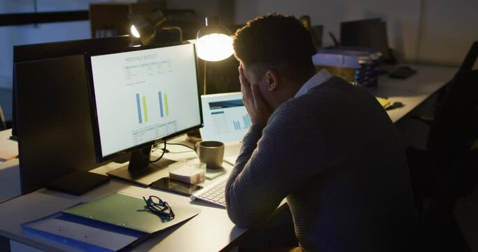 Video of tired biracial man using computer, working late in office