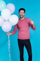 Positive Smiling Funny Caucasian Guy Handsome Brunet Man With Bunch of Colorful Air Balloons in Pink Jumper Showing Thumbs Up Against Blue Background.