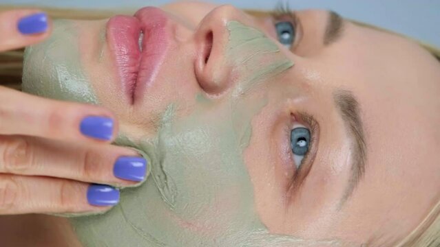 Close-up of the face of a young caucasian blonde woman. Green beauty mask made of clay on the face. Cosmetic procedure, rejuvenation and skin care. Blue background.