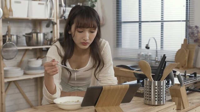asian girl leaning prone on kitchen table is selecting and watching videos on her touchpad while having a bowl of corn flake with milk for breakfast at home.
