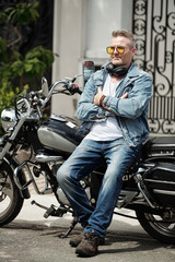 Fototapeta na wymiar Portrait of confident mature man in jeans and demin jacket leaning on motorcycle when waiting for girlfriend