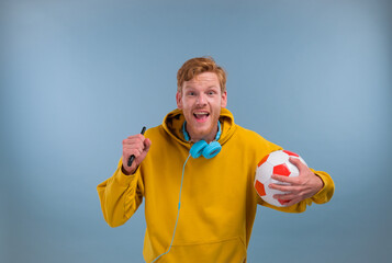 Photo of funny lucky young man wear purple hoodie smiling holding ball tv remote watching football match isolated on blue background - Powered by Adobe
