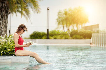Fit mature woman in red swuimsuit sitting on pool edge and reading captivating nodel - Powered by Adobe