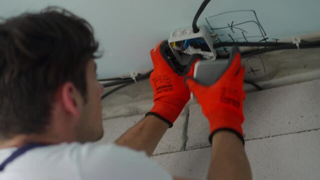 Electrician mounting junction box, instaling electrical wiring in apartment. Repairman feeds wires, cable into electrical box. Repair and construction work. Contractor working on apartment renovation.