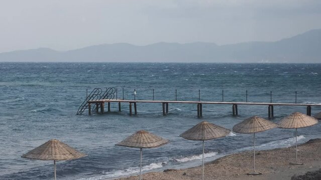 Parasol umbrellas on the beach bend in the wind. Old rusty pier swimming platform in Assos - Canakkale located at Aegean coasts of Turkey. Midilli - Lesbos Island in the background. Tilt view.