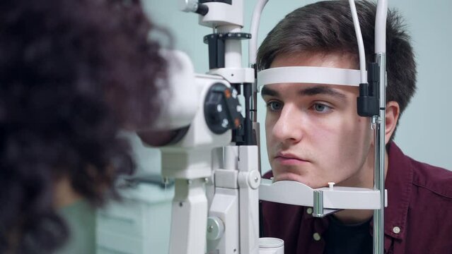 Portrait of concentrated man sitting at retina camera as woman checking back of the eye. Focused serious Caucasian patient consulting doctor ophthalmologist in hospital indoors