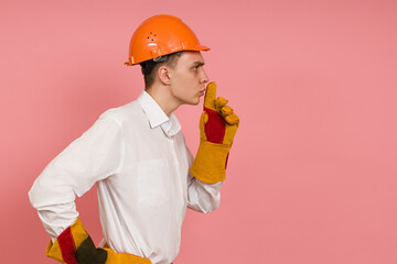 a young man in a white shirt, hard hat and protective gloves raised his finger to his mouth, stands sideways against a pink background with copy space