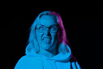 a woman in glasses in a hoodie made a face and shows her tongue in a blue neon light on a black background