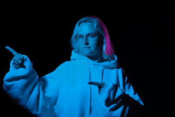 woman in glasses in a hoodie points her finger to the side in blue light on a black background