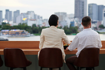 Fototapeta premium Business couple having cold beers and snacks and looking at beautiful cityscape front rooftop bar after work