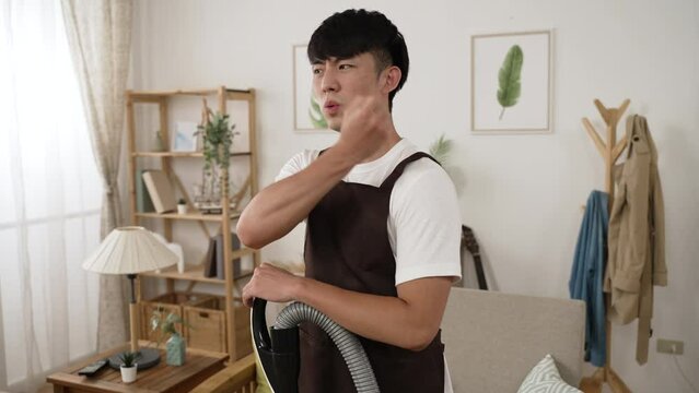 asian male cleaner having pain in his shoulders is exercising and pounding massaging the aching muscles with grimacing face while using hoover in the living room