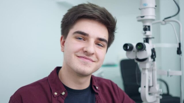 Young confident patient sitting at refractometer looking back at equipment turning smiling. Satisfied Caucasian man posing in ophthalmological clinic indoors. Optometry concept