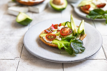 Bagels sandwich topped with cream cheese, farm cottage cheese, avocado, tomatoes, cucumbers and salad leaves, tilled background. Healthy breakfast food.