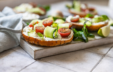 Bagels sandwich topped with cream cheese, farm cottage cheese, avocado, tomatoes, cucumbers and salad leaves, tilled background. Healthy breakfast food.