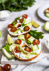 Bagels sandwich topped with cream cheese, farm cottage cheese, avocado, tomatoes, cucumbers and salad leaves, tilled background. Healthy breakfast food.