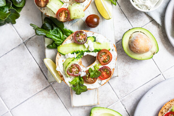Bagels sandwich topped with cream cheese, farm cottage cheese, avocado, tomatoes, cucumbers and salad leaves, tilled background. Healthy breakfast food.