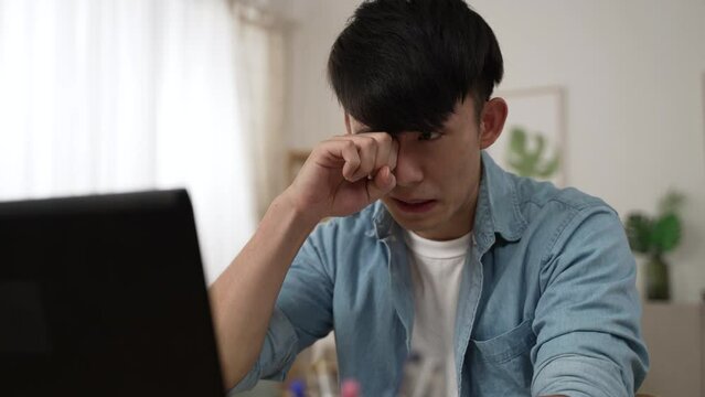 shoulder shot of an overworked asian male employee rubbing his tired eyes while working from home on the laptop in the living room at daytime.