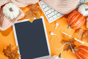 Cozy fall background with tablet, ear-pods, white, orange pumpkins, autumn leaves decor on high-colored orange background. Autumn still life composition.