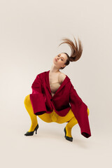 Portrait of young girl in yellow tights, red jacket and heel shoes, sitting on haunches, posing isolated over grey studio background