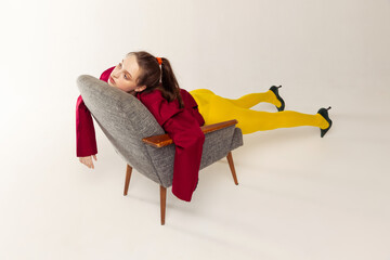 Portrait of young girl in yellow tights and red jacket posing with chair isolated over grey studio...