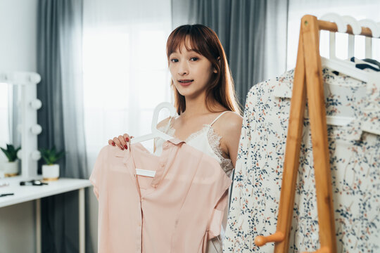 portrait asian woman holding a blouse on the hanger is looking at her reflection in the mirror while changing clothes by a clothing rack in a bright bedroom.