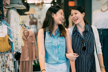 portrait two asian sister besties are smiling at each other while chatting and shopping arm in arm in a woman’s clothing shop